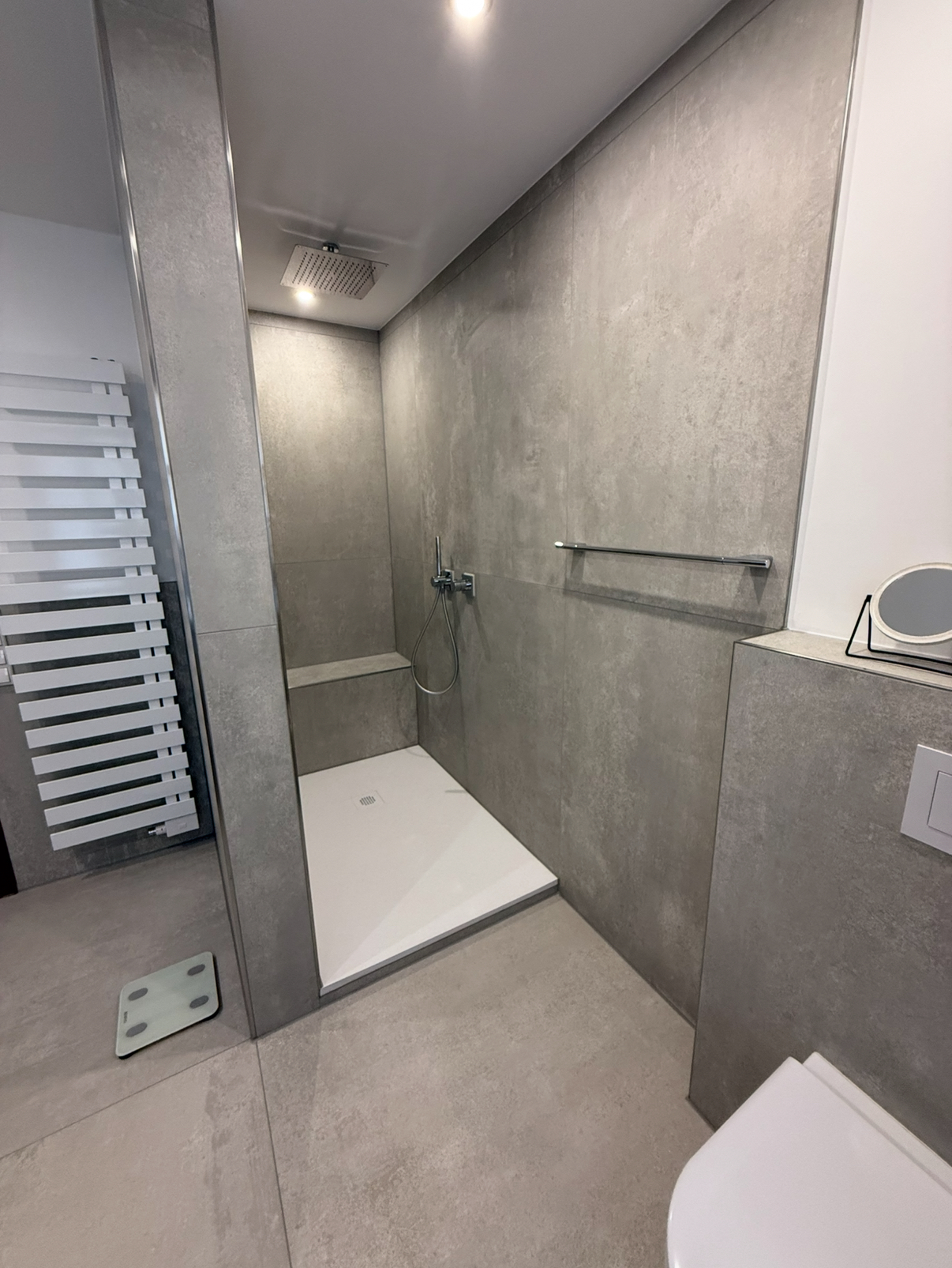 Moderner Duschbereich mit bodengleicher Duschwanne, grauen Wand- und Bodenfliesen, Handtuchhalter und Toilette im Vordergrund.