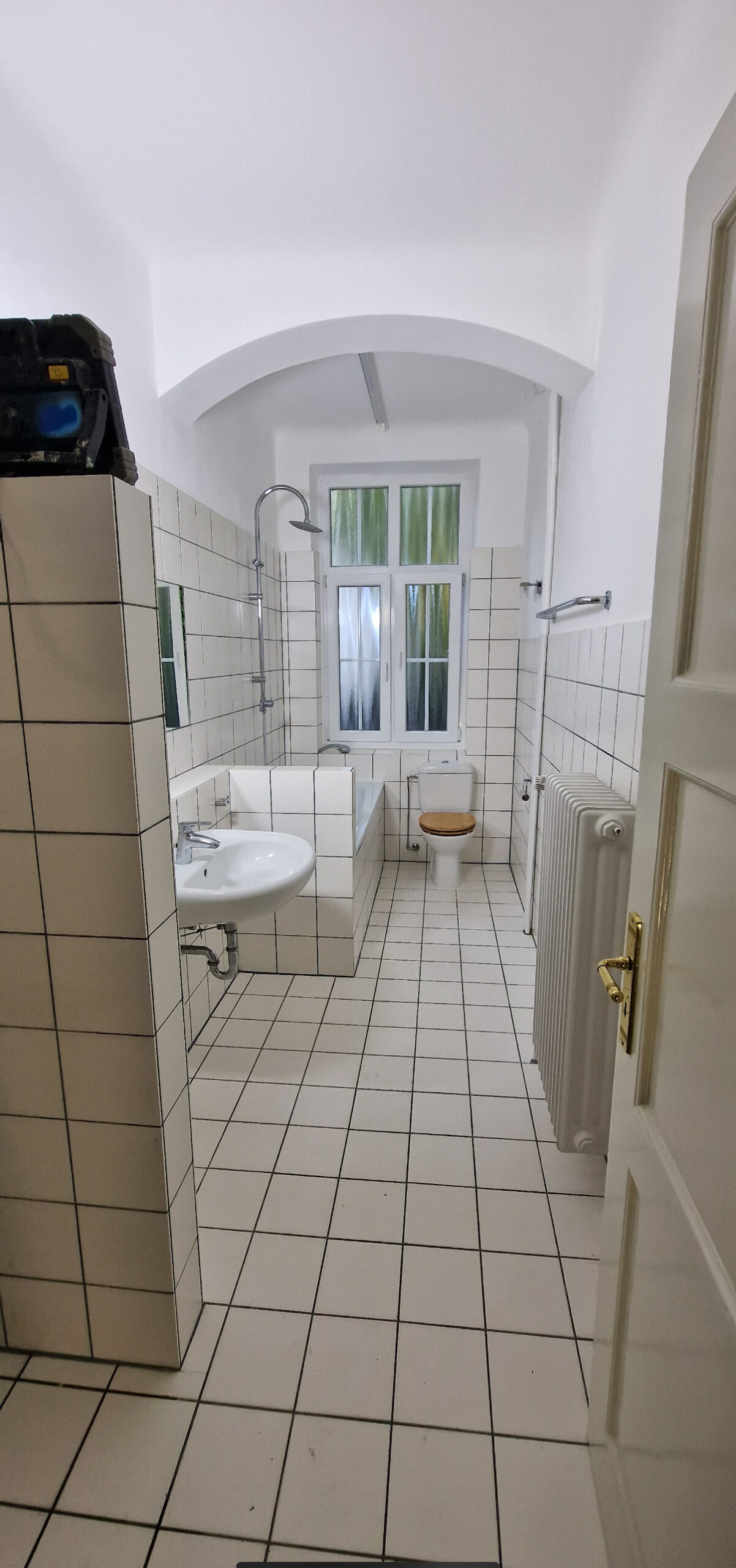 Schmaler, langgestreckter Raum mit weißen Fliesen an Boden und Wänden, Waschbecken, Badewanne, Toilette und Fenster am Ende.