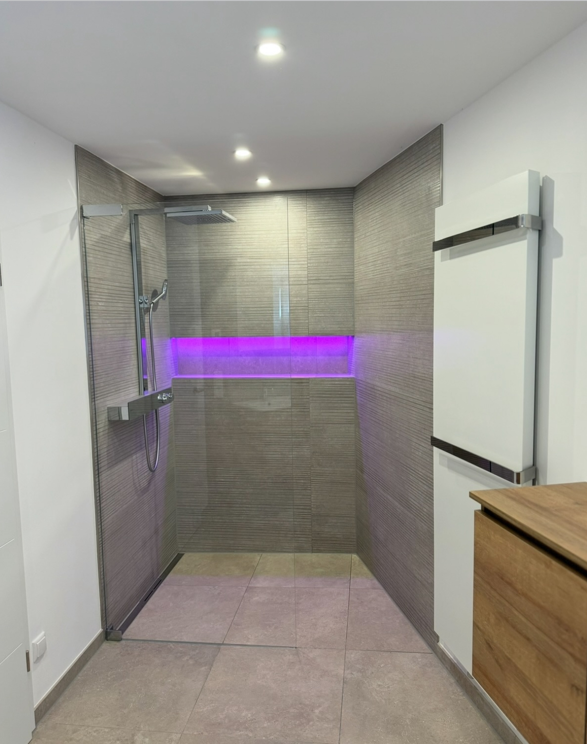 Moderne Dusche mit grauen Wandfliesen, bodengleicher Duschbereich, Glasabtrennung und lila LED-Beleuchtung in der Nische.
