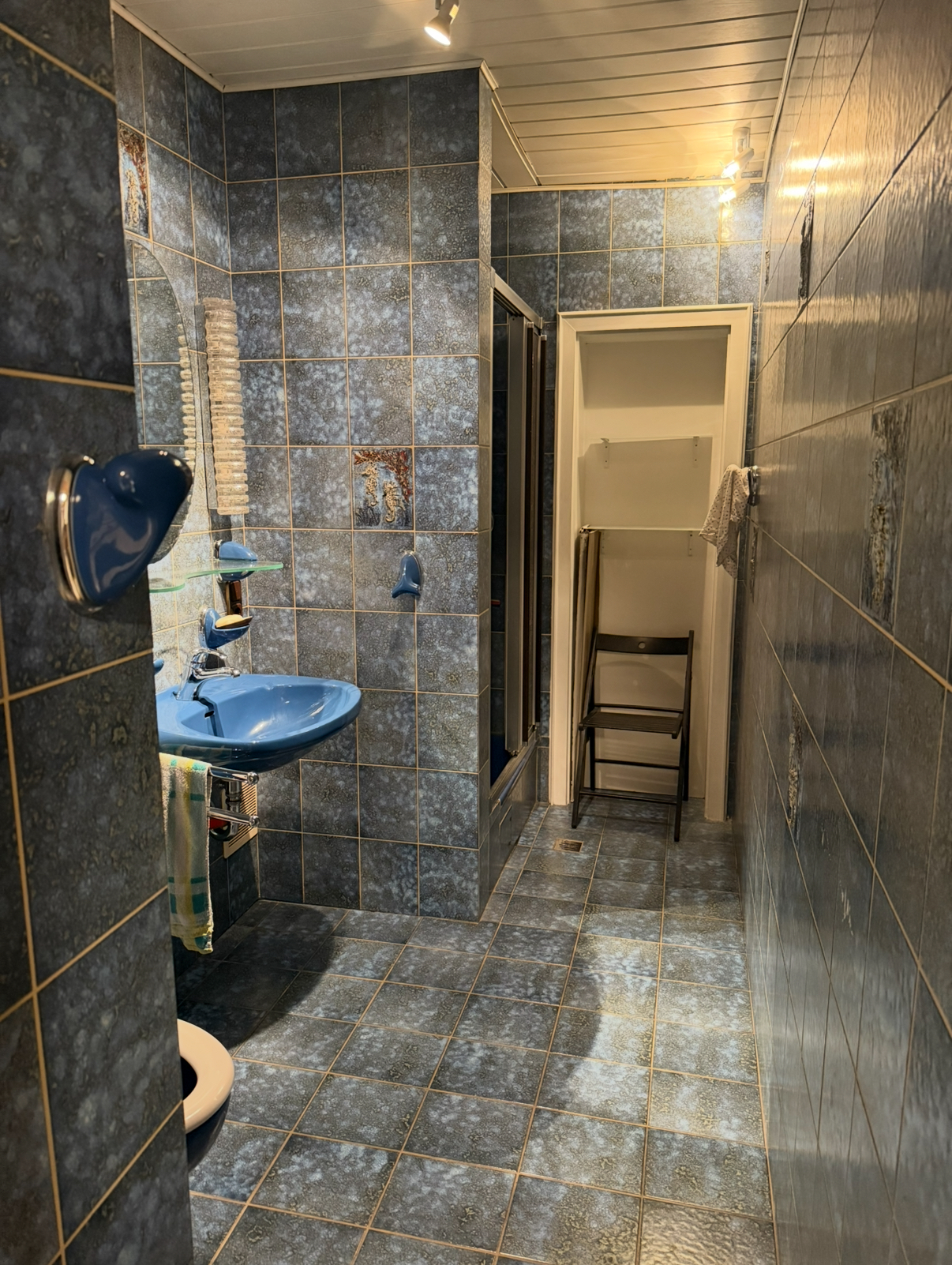 Schmaler, langgestreckter Flur mit blau-grauen Fliesen an Boden und Wänden, blauem Waschbecken und Duschkabine am linken Rand, am Ende ein offener Durchgang mit Klappstuhl davor.