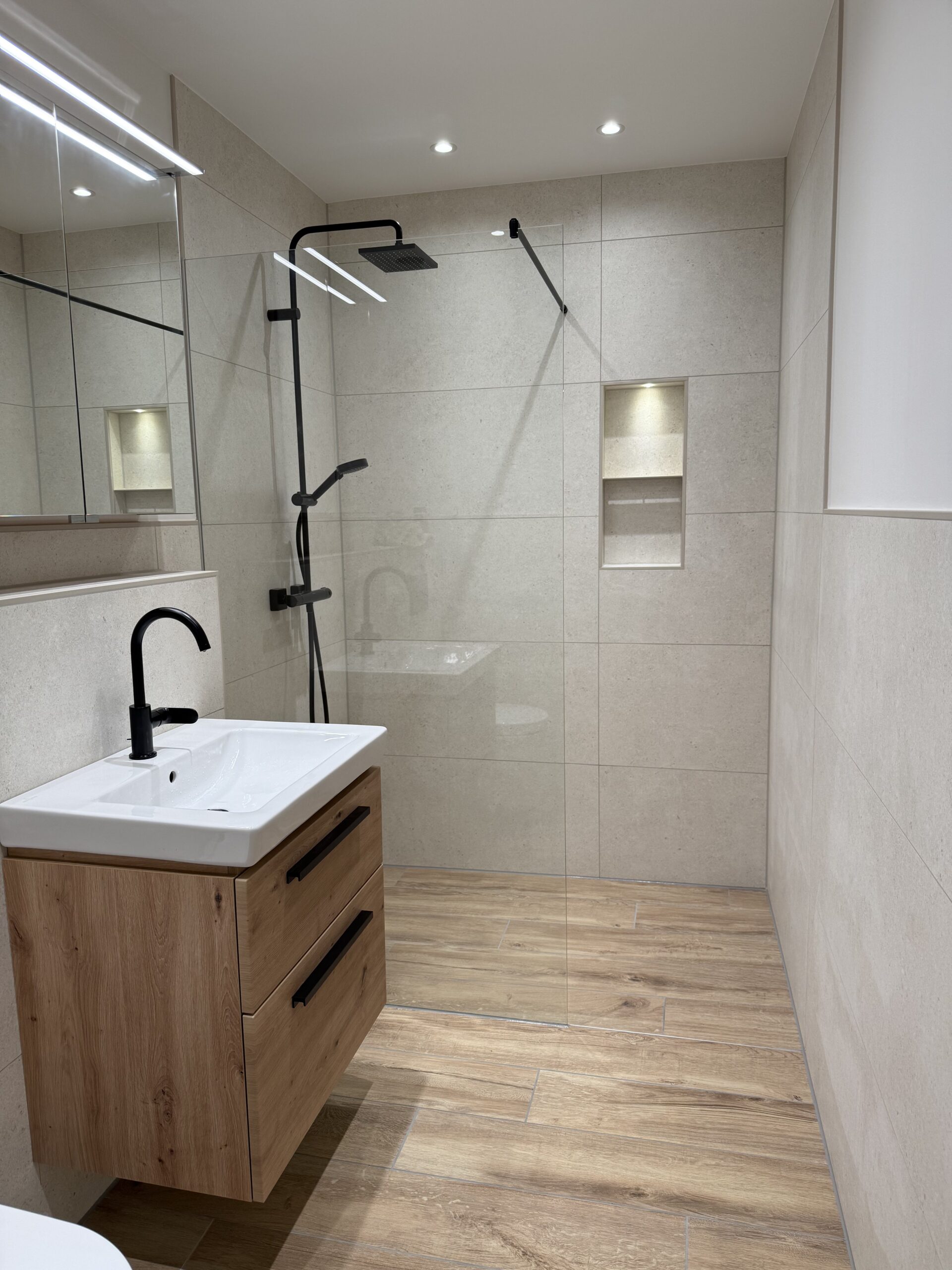 Modernes Badezimmer mit bodengleicher Dusche, Glasabtrennung, Holzoptik-Bodenfliesen und Waschtisch mit schwarzem Wasserhahn und Holzschrank.