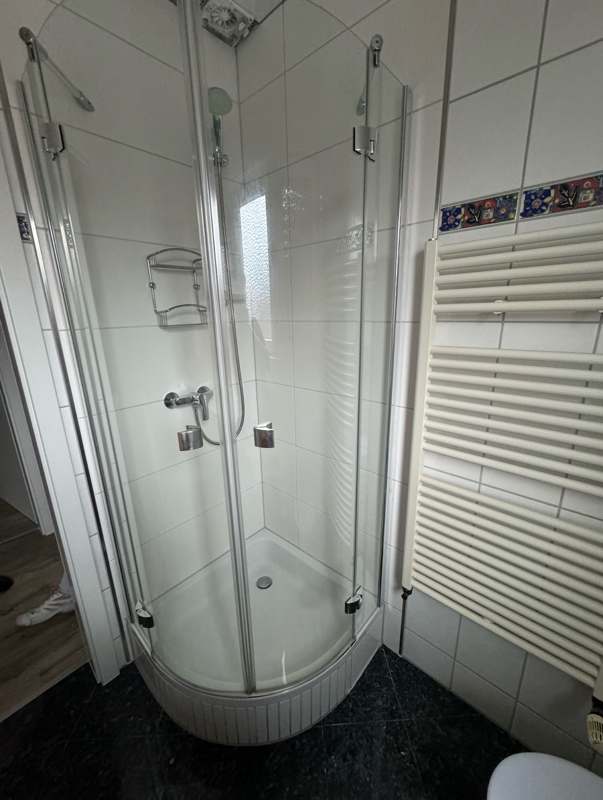 Eckdusche mit halbkreisförmiger Duschwanne, Glasfaltwand und verchromtem Duschkopf in einem gefliesten Badezimmer.