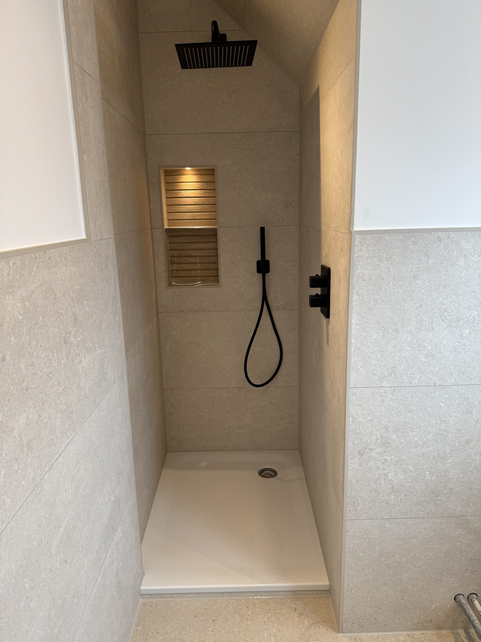 Schlichte Dusche mit quadratischem Regenduschkopf, schwarzem Handbrausehalter und Ablagefach in beige gefliester Nische.